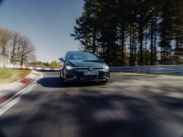 A green VW Golf GTi Edition 50 speeds around a race track with red-and-white barriers and a forest backdrop; motion blur emphasises speed.