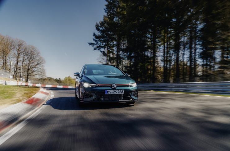 A green VW Golf GTi Edition 50 speeds around a race track with red-and-white barriers and a forest backdrop; motion blur emphasises speed.