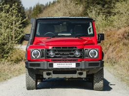 Front view of a red SUV with a black grille, round headlights, and a white license plate reading 'GRENADIER TRIAL MASTER X' on a gravel road.