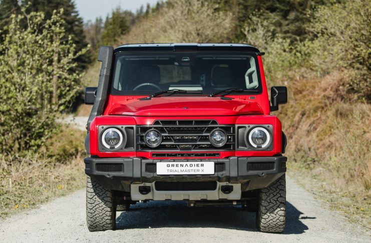 Front view of a red SUV with a black grille, round headlights, and a white license plate reading 'GRENADIER TRIAL MASTER X' on a gravel road.