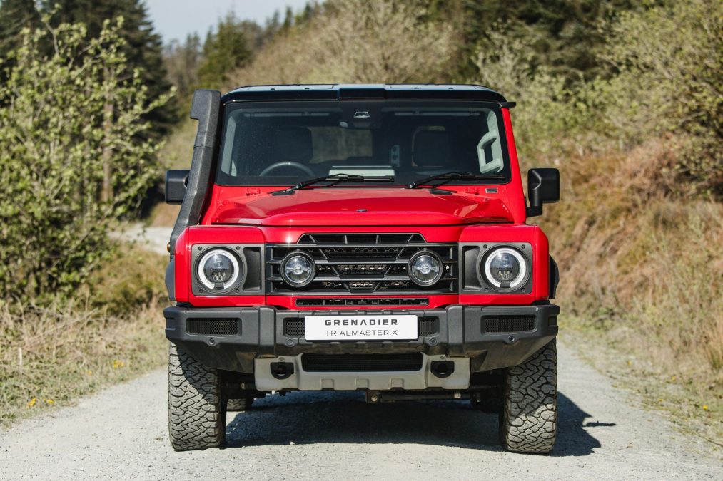 INEOS Grenadier Trialmaster X Front view of a red SUV with a black grille, round headlights, and a white license plate reading 'GRENADIER TRIAL MASTER X' on a gravel road.