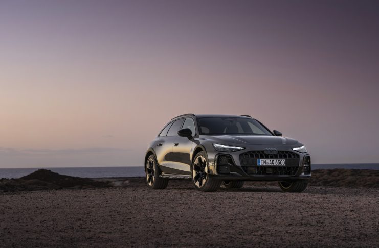 Silver Audi SUV parked on a rocky coastal shore at sunset with calm water in the background.