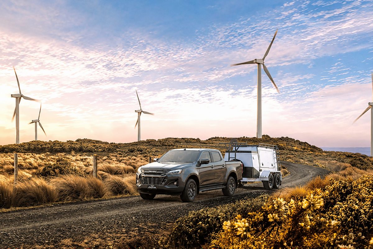 Gray pickup truck towing a silver box trailer along a dusty rural road with wind turbines and desert scrub under a pastel sunset.