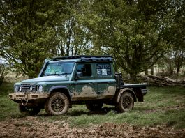 Green 4x4 SUV with a roof rack parked in a muddy field, with dense trees in the background.