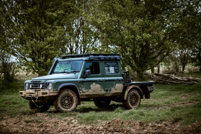 Green 4x4 SUV with a roof rack parked in a muddy field, with dense trees in the background.